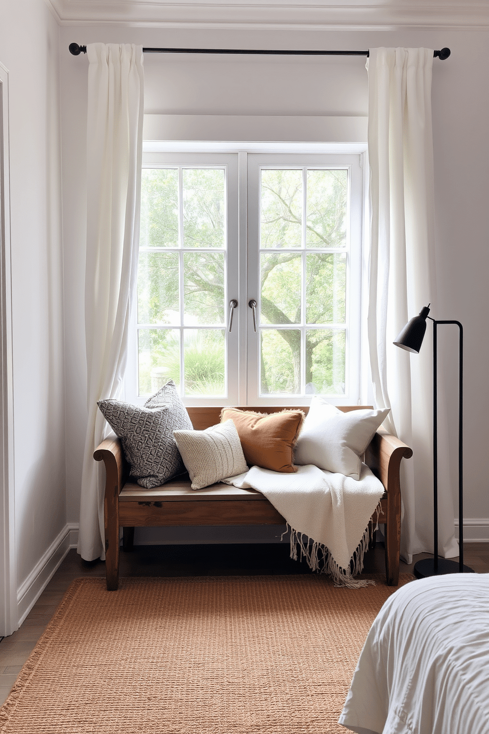 A cozy reading nook in a serene bedroom setting. A rustic wooden bench with a distressed finish is set beneath a large picture window, framed by soft white linen curtains that pool slightly on the floor. The bench is layered with a plush cream throw blanket and an assortment of textured pillows in warm earth tones. A woven jute rug anchors the space, while a slender black floor lamp with a minimalist design stands nearby, casting a soft golden glow over the corner.