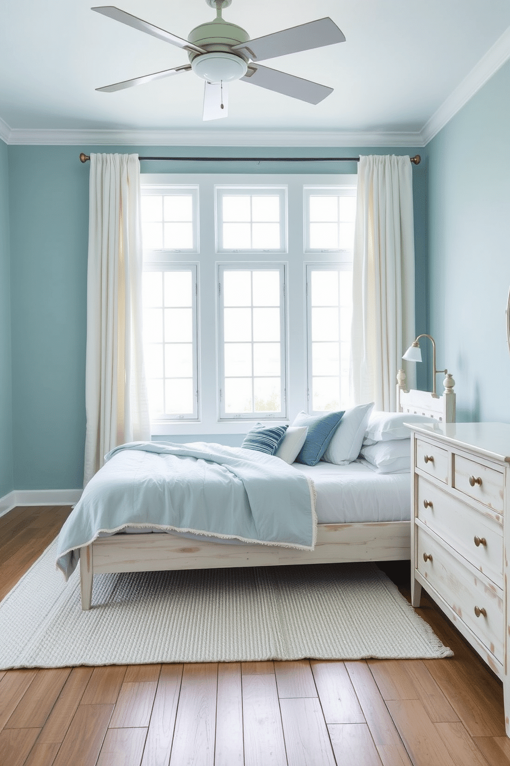A serene bedroom radiating coastal charm. The walls are painted in a soft aqua blue, complemented by whitewashed wooden furniture, including a low-profile bed and a matching dresser adorned with minimal brass hardware. A textured white rug lies beneath the bed, while airy linen curtains frame large windows that allow natural light to flood the space. Decorative pillows in varying shades of blue and white rest on the bed, paired with a lightweight quilt that furthers the breezy, ocean-inspired ambiance.