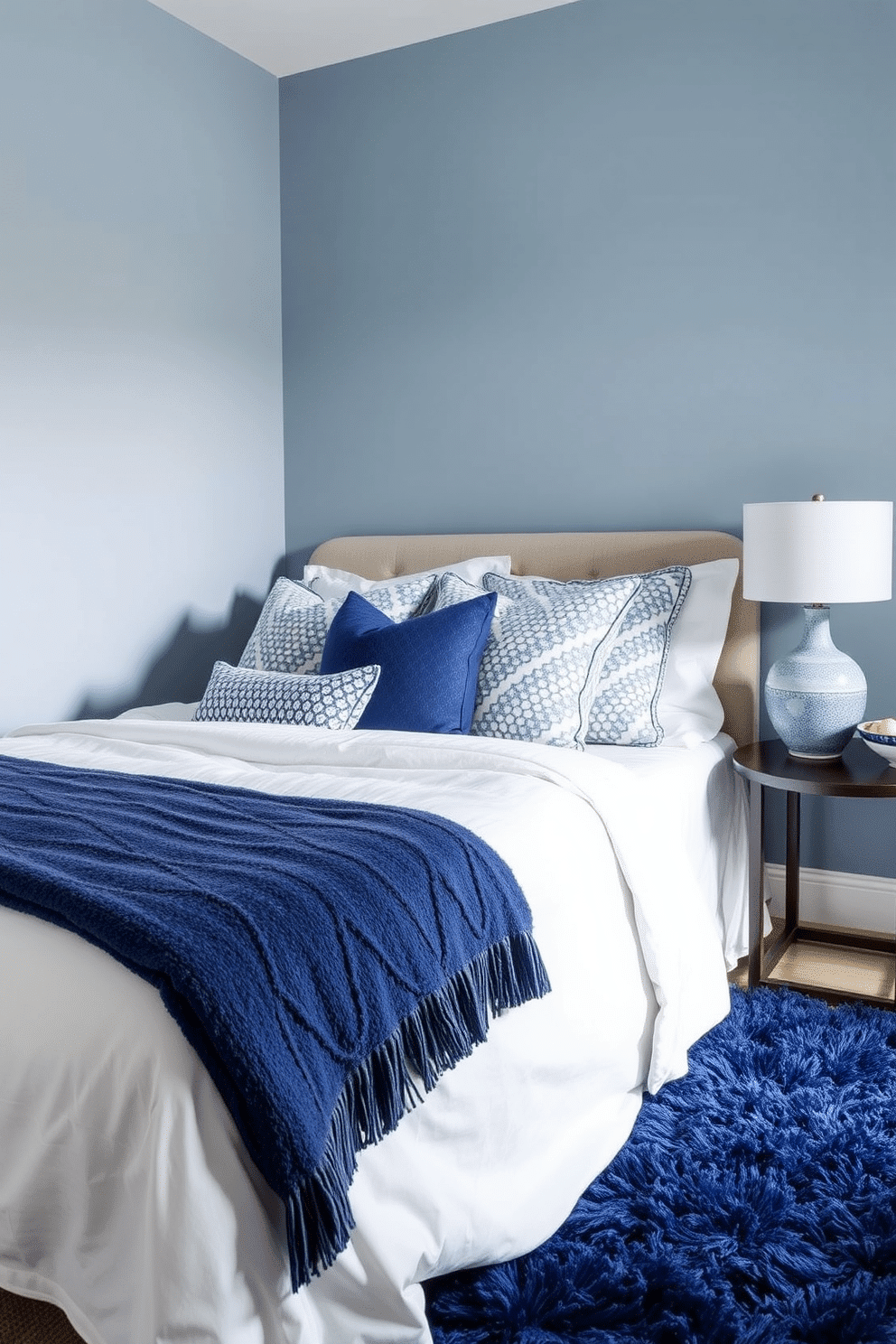 A serene bedroom with walls painted in a soft, dusty blue hue to create a calming atmosphere. The bed is adorned with crisp white linens and accented with an assortment of blue patterned throw pillows, adding layers of texture and visual interest. A plush navy area rug anchors the room, complementing the lighter blues and offering warmth underfoot. On the bedside table, a ceramic lamp with a textured blue base sits beside a small decorative bowl, tying the palette together seamlessly.
