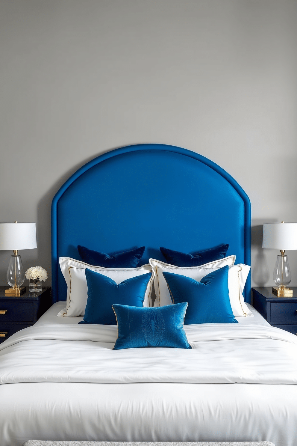 A luxurious bedroom with a striking peacock blue arched statement headboard as the focal point. The bed is dressed in crisp white linens with subtle gold accents, complemented by plush throw pillows in varying shades of blue and teal. The walls are painted in a soft dove gray, creating a sophisticated contrast with the vibrant headboard. On either side, sleek navy bedside tables with brass hardware hold modern glass lamps with white shades, casting a warm glow over the serene space.
