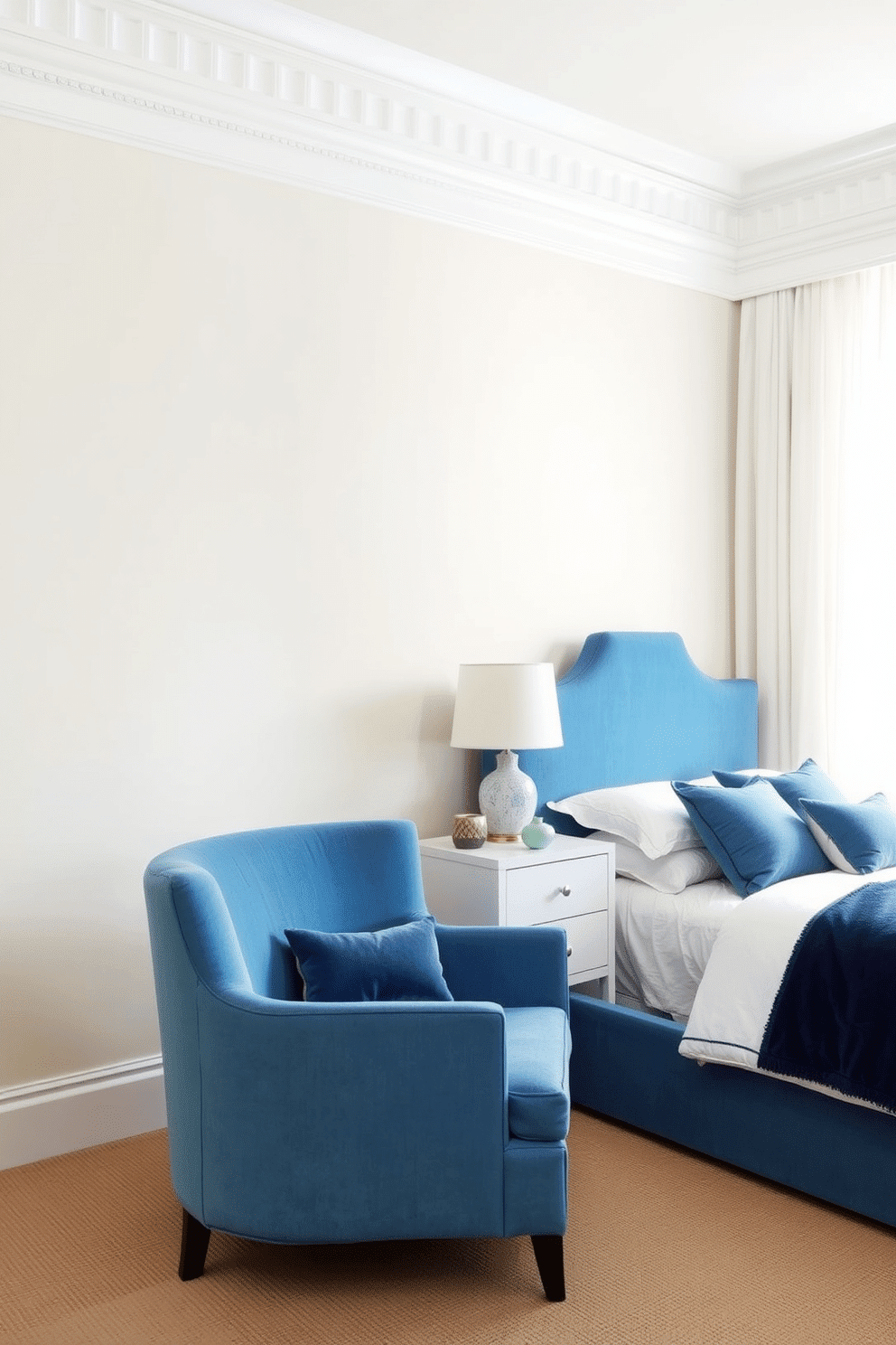 A serene bedroom with pale, neutral-colored walls that create a soft and calming backdrop. Cornflower blue upholstered chairs sit beside a sleek, white bedside table, complementing the plush blue headboard of the bed. The bedding features layered blue and white linens, creating a harmonious look with the furniture. Sheer white curtains allow natural light to filter gently into the space, highlighting decorative ceramic vases and a stylish table lamp in muted blue tones.