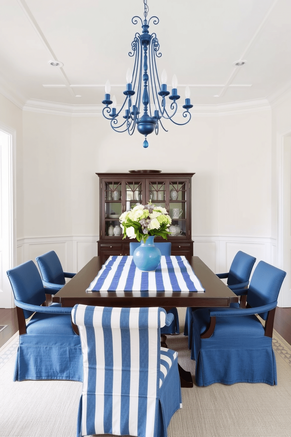 A stylish dining room featuring blue and white striped table linens draped elegantly over a dark wooden table. Surrounding the table are upholstered chairs in a complementary solid blue fabric, creating a cohesive and inviting atmosphere. The walls are painted in a soft white tone, enhancing the brightness of the space, while a statement chandelier with blue accents hangs above the table. A centerpiece of fresh flowers in a blue vase adds a touch of color and vibrancy to the setting.