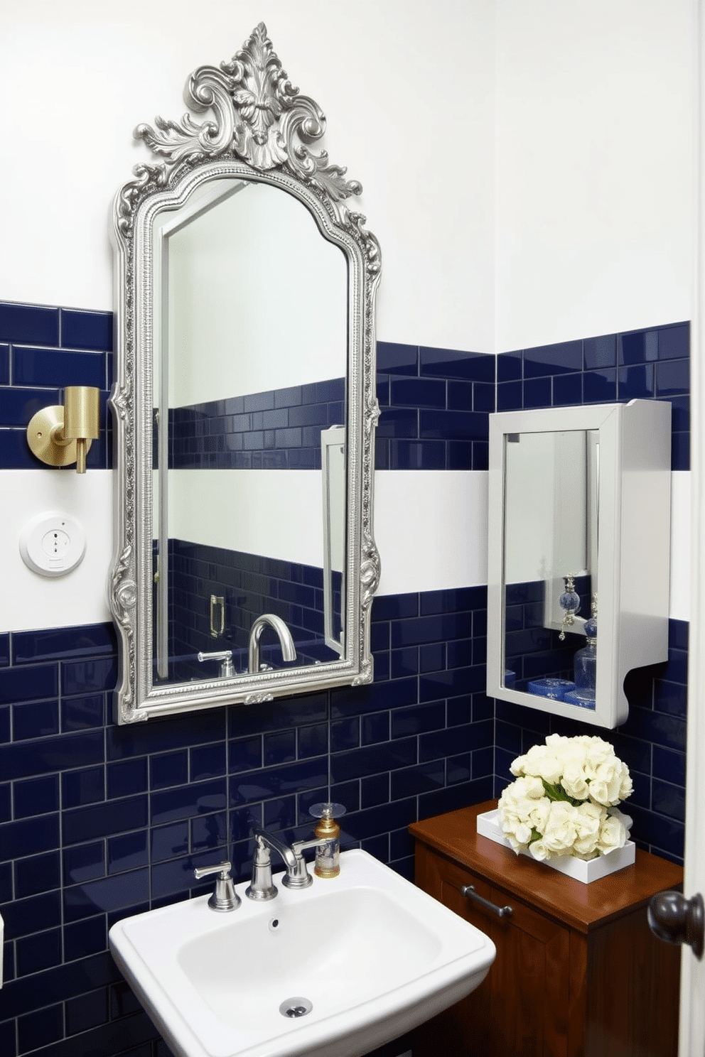 A chic powder room featuring navy blue tiles elegantly framing a large, ornate mirror. The walls are painted in a soft white, creating a striking contrast with the deep blue tiles, and a sleek pedestal sink complements the design. To the side, a small wooden cabinet provides storage while maintaining an airy feel. Decorative accents, such as a stylish soap dispenser and a fresh bouquet of white flowers, enhance the room's sophistication.