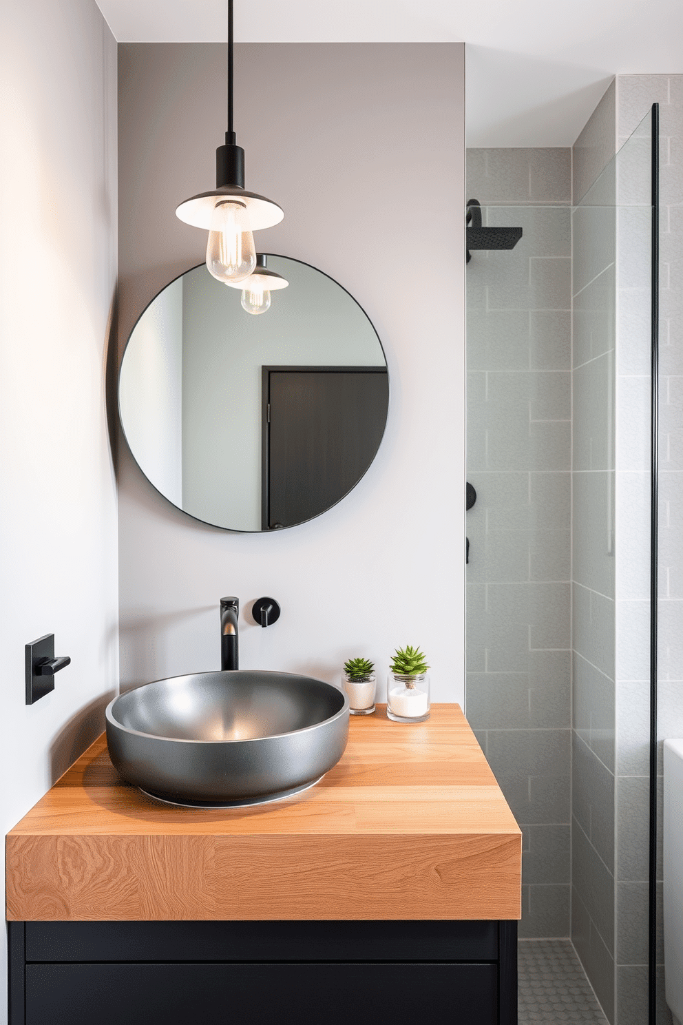 A contemporary bathroom design featuring a steel-gray sink seamlessly integrated into a natural wooden countertop. Above, a frameless round mirror reflects light from a modern pendant fixture, casting a warm glow. The walls are painted in a soft dove-gray, complemented by textured subway tiles in the shower area. A small potted plant and neatly rolled towels add subtle greenery and softness to the space.