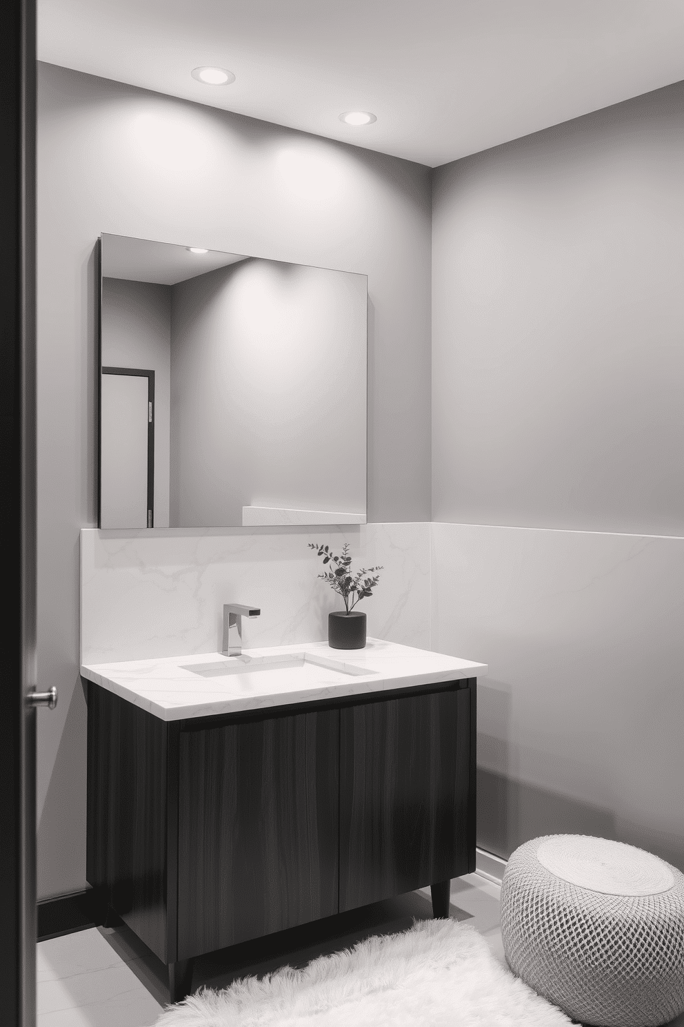 A sleek and modern bathroom setting with light gray marble adorning both the countertop and backsplash, exuding sophistication. The vanity features clean-lined wooden cabinetry in a matte black finish, complemented by brushed nickel hardware for a contemporary touch. Above the countertop, a frameless mirror stretches across the wall, reflecting soft, ambient lighting from recessed fixtures in the ceiling. The muted gray walls pair harmoniously with the marble details, while a plush white rug and a small potted plant add texture and warmth to the space.