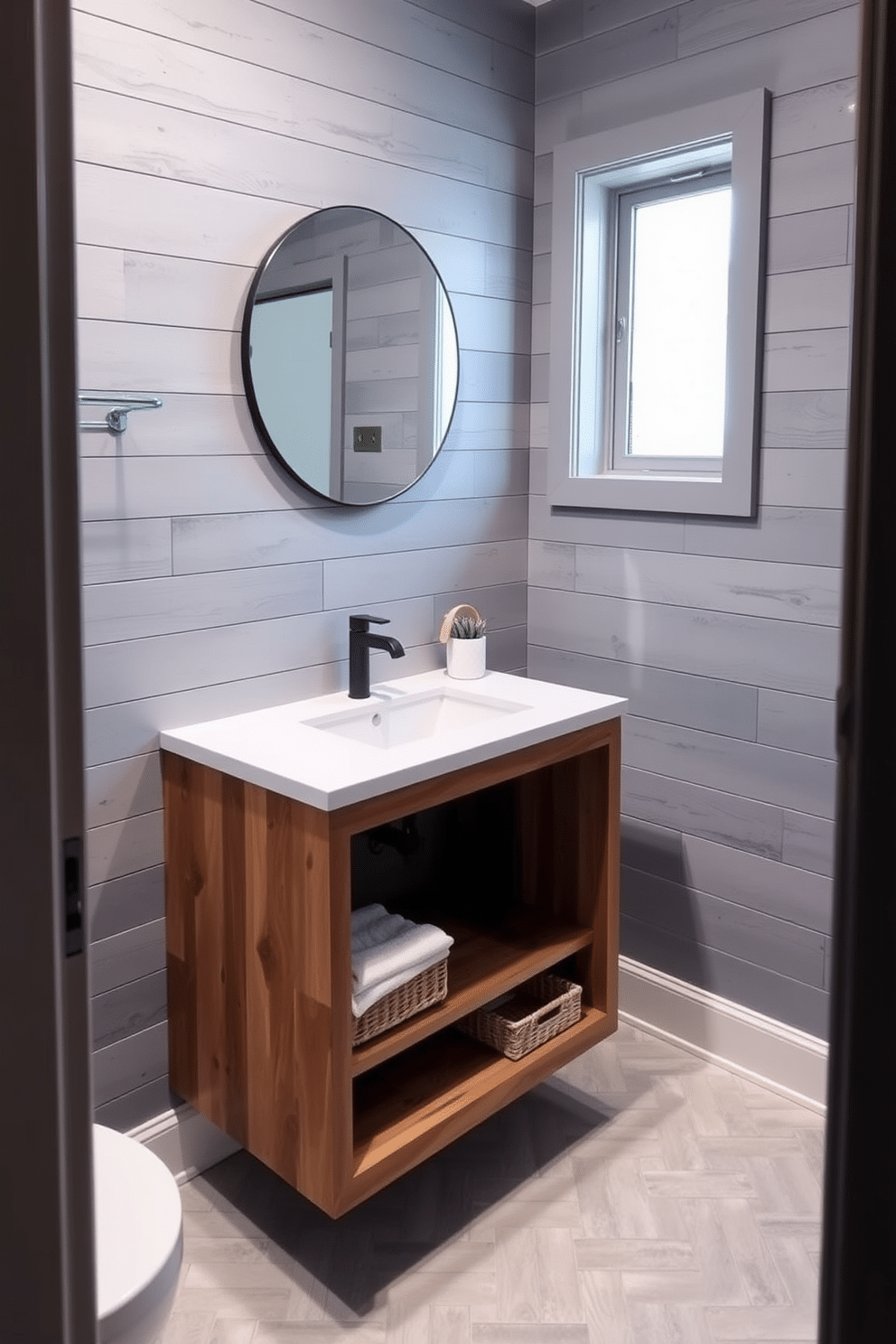 A cozy yet modern bathroom space with a gray shiplap accent wall as the backdrop, adding a rustic charm to the room. The shiplap wall complements a floating wooden vanity with a white quartz countertop, featuring a sleek undermount sink and matte black fixtures. Above the vanity, a round, frameless mirror reflects soft natural light streaming in from a small frosted window. The flooring showcases light gray herringbone tiles, while a woven basket and a folded white towel rest on the open shelf of the vanity for added texture.