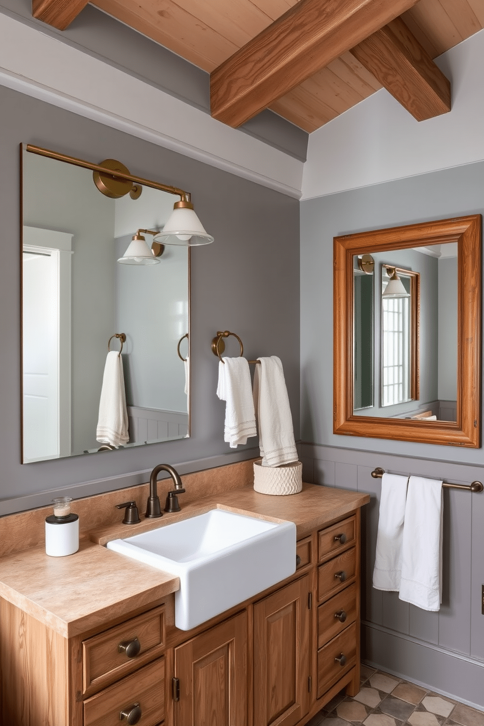 A cozy rustic farmhouse bathroom with walls painted in two-toned shades of gray, combining a lighter hue on top with a deeper shade on the bottom. Exposed wooden beams on the ceiling and aged brass fixtures add warmth to the space. The vanity features weathered wood cabinetry with a natural stone countertop and a deep farmhouse sink. Large framed mirrors, pendant lighting, and soft linen accents in white and cream complete the inviting yet sophisticated look.