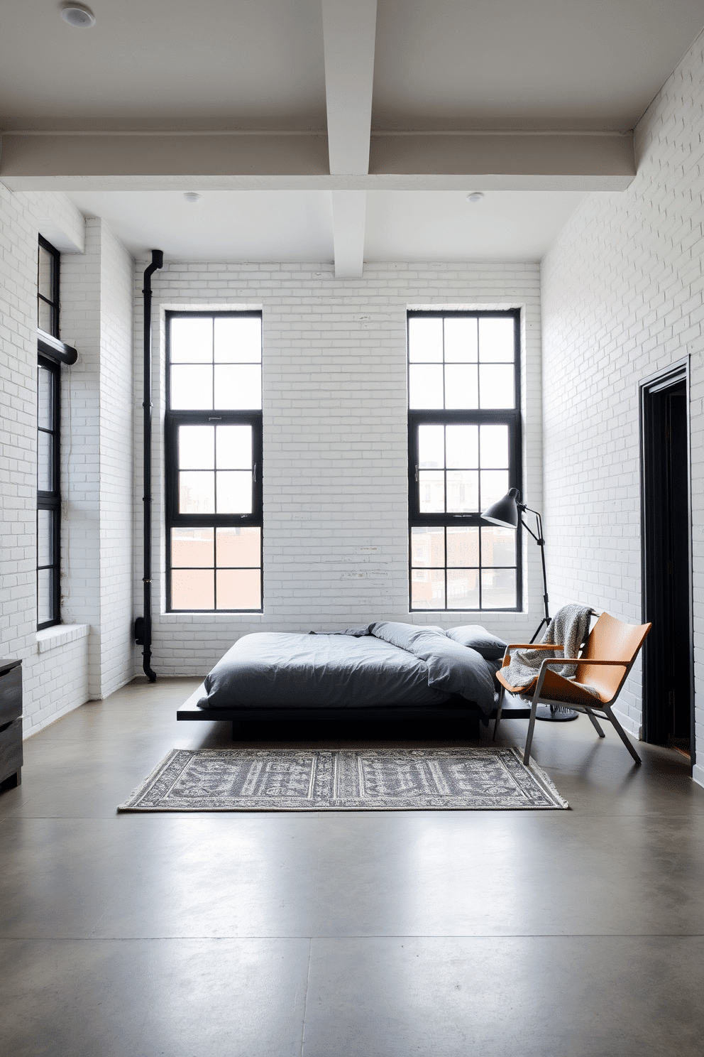 A contemporary loft bedroom with painted white brick walls for a modern, textured aesthetic. A low platform bed with minimalist gray bedding sits in the center of the space, complemented by black metal-framed windows flooding the room with natural light. In one corner, a sleek wooden armchair is paired with a cozy throw blanket, near a tall industrial floor lamp for added warmth. The floors are polished concrete, and a geometric area rug adds a subtle pop of pattern, completing the urban-chic atmosphere.