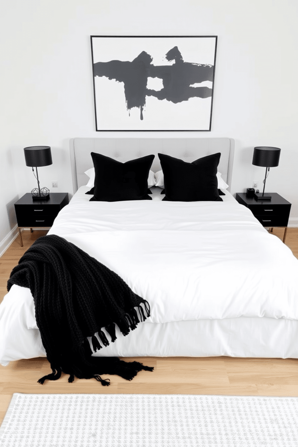 A minimalist bedroom with clean white bedding accented by black throw pillows and a matching black knit blanket folded at the foot of the bed. The headboard is an upholstered panel in soft, neutral fabric, framed by sleek black bedside tables with simple, geometric lamps. The walls are painted bright white to complement the airy and uncluttered aesthetic, while a large black-and-white abstract piece of art hangs above the bed. Beneath, a low-profile modern rug in soft gray tones contrasts gently against light oak wood floors for added warmth and sophistication.