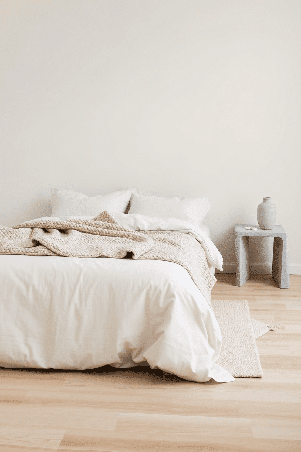 A serene minimalist bedroom with an emphasis on textures. The bed is dressed in soft linen bedding, paired with a chunky knit throw laid across the end. The floor showcases light natural wood planks, while the walls are smooth and painted in a warm, matte off-white. A neutral, low-pile area rug adds subtle coziness underfoot, and a sleek stone nightstand holds a single ceramic vase.
