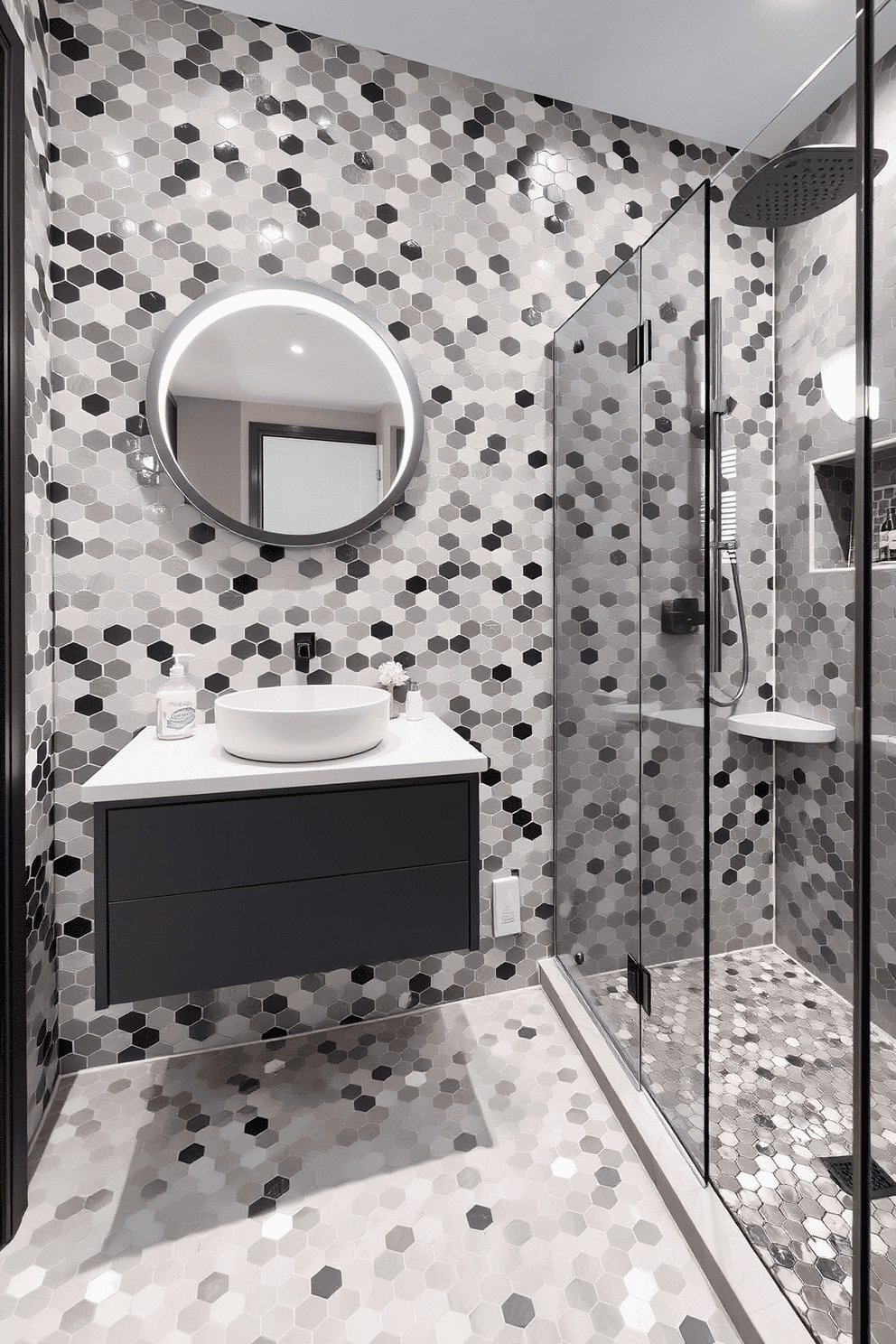 A contemporary bathroom showcasing hexagonal mosaic tiles in varying shades of gray, creating a visually dynamic statement wall behind a sleek floating vanity. The vanity is topped with a white quartz countertop holding a round ceramic sink, while a circular backlit mirror above adds a subtle modern glow. The floor continues the hexagonal theme with matching mosaic tiles, seamlessly blending into the overall design. A glass-enclosed walk-in shower features a rainfall showerhead, with a recessed shelf lined with contrasting accent tiles for added texture and interest.