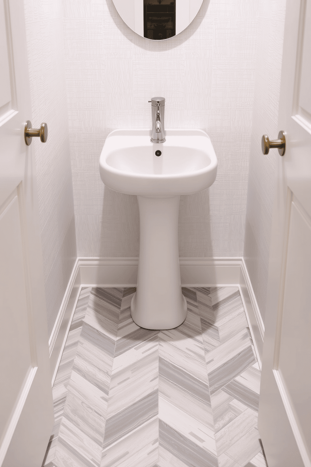 A chic powder room featuring a herringbone pattern floor tile that creates a striking visual interest. The tiles are in a mix of soft gray and white, adding a touch of elegance to the compact space. The walls are adorned with a subtle textured wallpaper in a pale hue, complementing the floor design. A sleek, modern pedestal sink is centered against the wall, accentuated by a stylish wall-mounted faucet in brushed nickel.