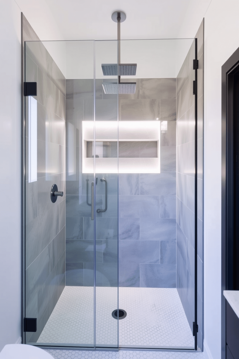 An open shower layout featuring a frameless glass partition that seamlessly blends into the bathroom's minimalist aesthetic. The shower walls are clad in large-format matte gray tiles, while the floor contrasts with small hexagonal white tiles for added texture. A sleek rain showerhead is mounted on the ceiling, flanked by a recessed shelf for toiletries. Soft, diffused LED lighting illuminates the shower space, adding a warm ambiance, while the rest of the bathroom showcases clean lines and modern finishes.