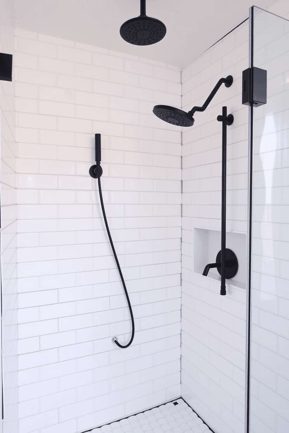 A sleek walk-in shower featuring clean white subway tiles arranged in a classic brick pattern. The shower fixtures, including a rain showerhead and a hand shower, are matte black, creating a striking contrast against the bright tiles. A frameless glass shower enclosure enhances the minimalist and contemporary aesthetic. A recessed shelf, also adorned with white tiles, provides storage, while a subtle black grout detail ties the design together seamlessly.