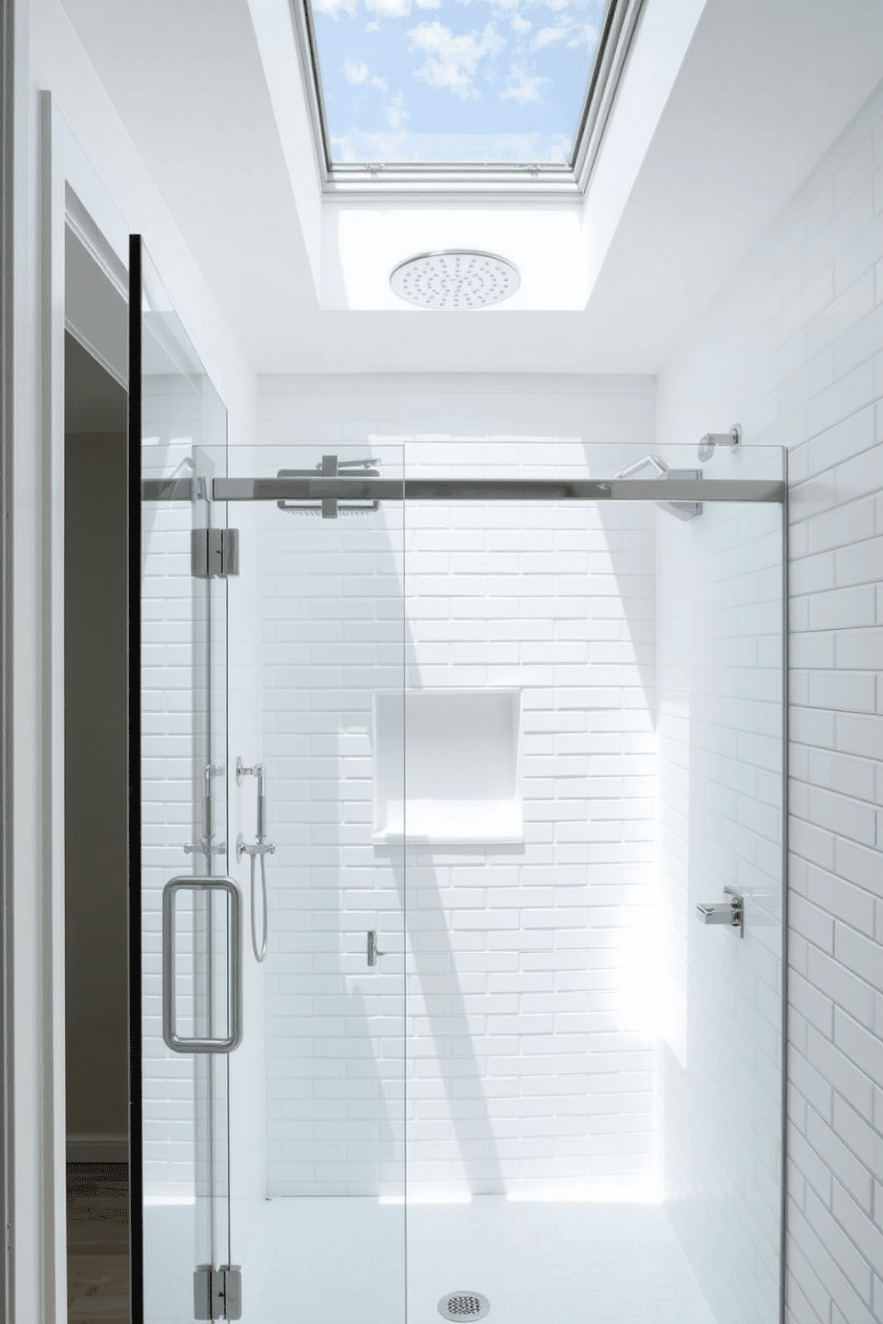 An ethereal walk-in shower with an all-white aesthetic, exuding simplicity and sophistication. The walls are adorned with smooth white subway tiles while a frameless glass door allows for a seamless flow of light. Above, a skylight invites natural sunlight to cascade into the space, creating a serene, spa-like atmosphere. A sleek chrome rain shower head and built-in white niche enhance the shower's functionality and minimalist charm.